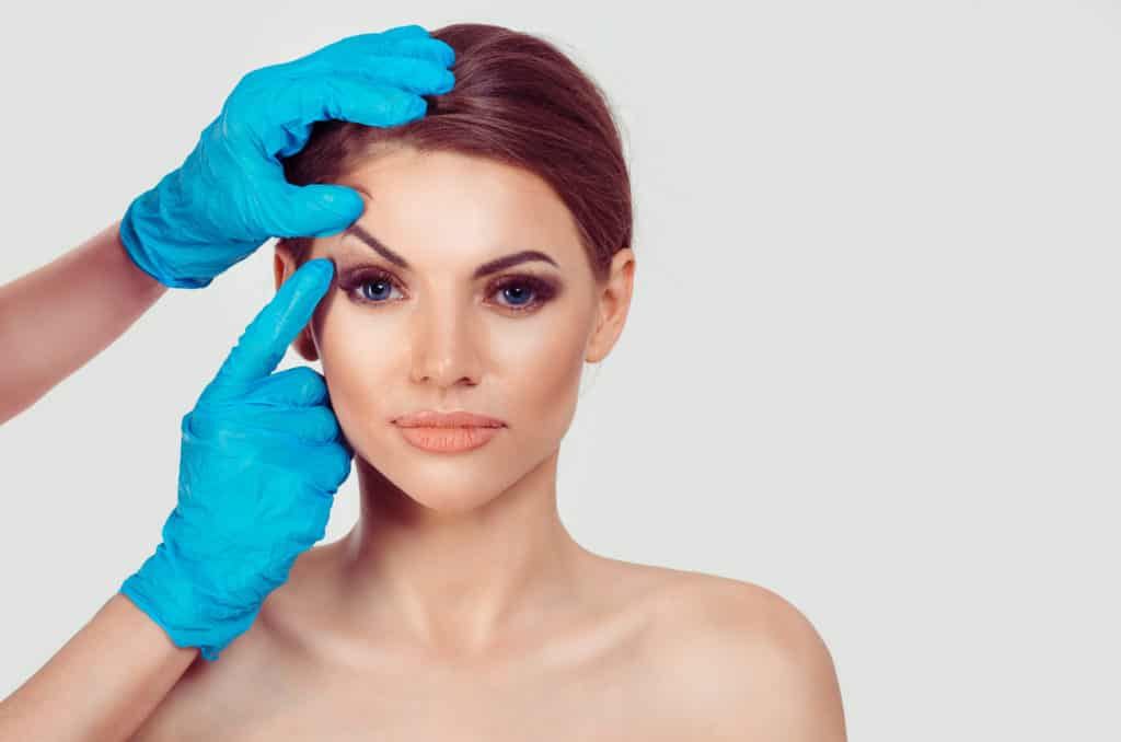 Upper eyelid blepharoplasty. Beautiful middle age woman getting ready for eyelid lift plastic surgery doctor hands in blue gloves point fingers to her eye on white. Beauty, people and health concept | Adam J Cohen, MD in Glenview & Chicago, IL