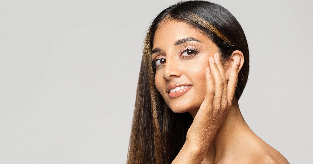 Studio portrait of a smiling young woman with highlighted dark hair, bare shoulders, and her hand on her cheek, representing Full Facelift in Glenview, IL, against a light gray background.